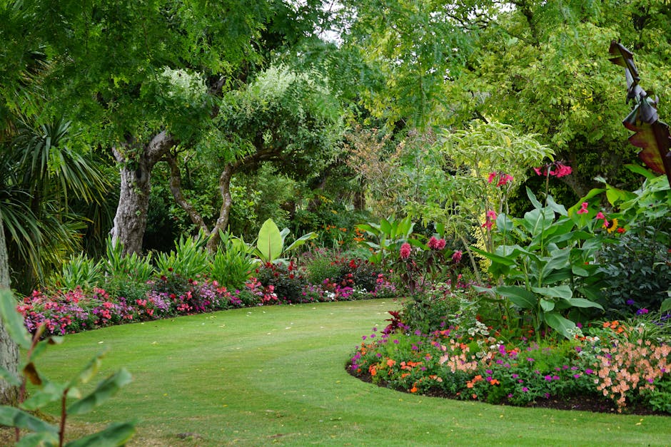 A lush garden scene featuring a well-maintained, vibrant green lawn edged with colorful flower beds containing pink, red, purple, and orange blossoms. Mature trees with gnarled trunks and dense, leafy