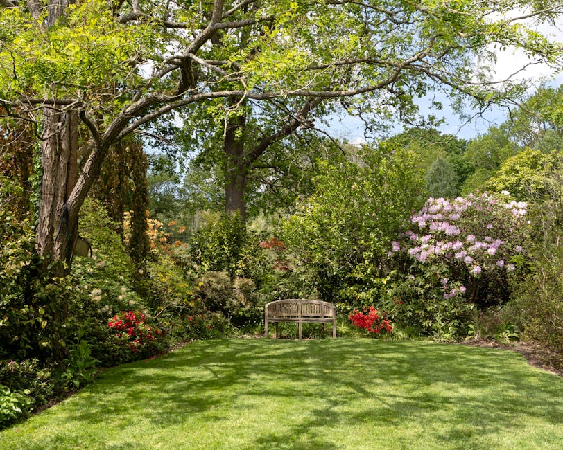 A lush garden scene featuring a well-maintained, vibrant green lawn edged with colorful flower beds containing pink, red, purple, and orange blossoms. Mature trees with gnarled trunks and dense, leafy