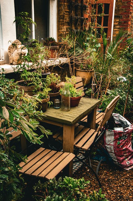 A cozy garden patio scene featuring a weathered wooden table surrounded by matching wooden benches and chairs, set against a backdrop of a brick house with a white-framed window and a wooden door. The