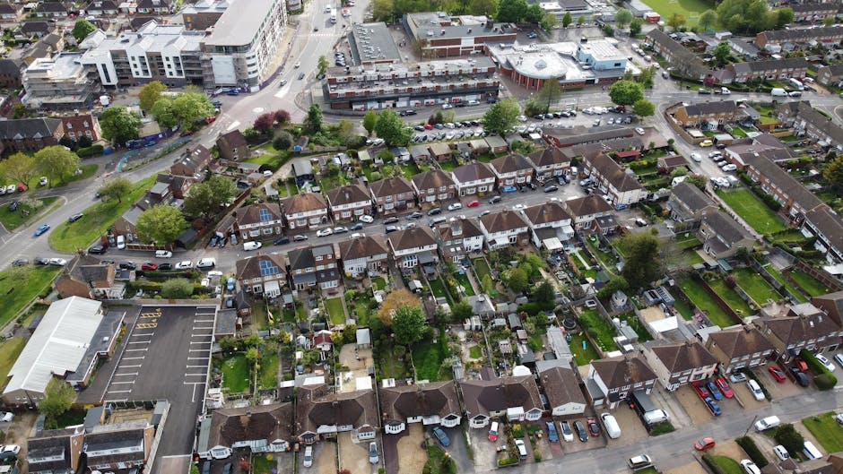 An aerial view of a suburban neighborhood showing a mix of residential houses, some with small gardens and sheds at the back, and surrounding green lawns, trees, and pathways. The houses are arranged 